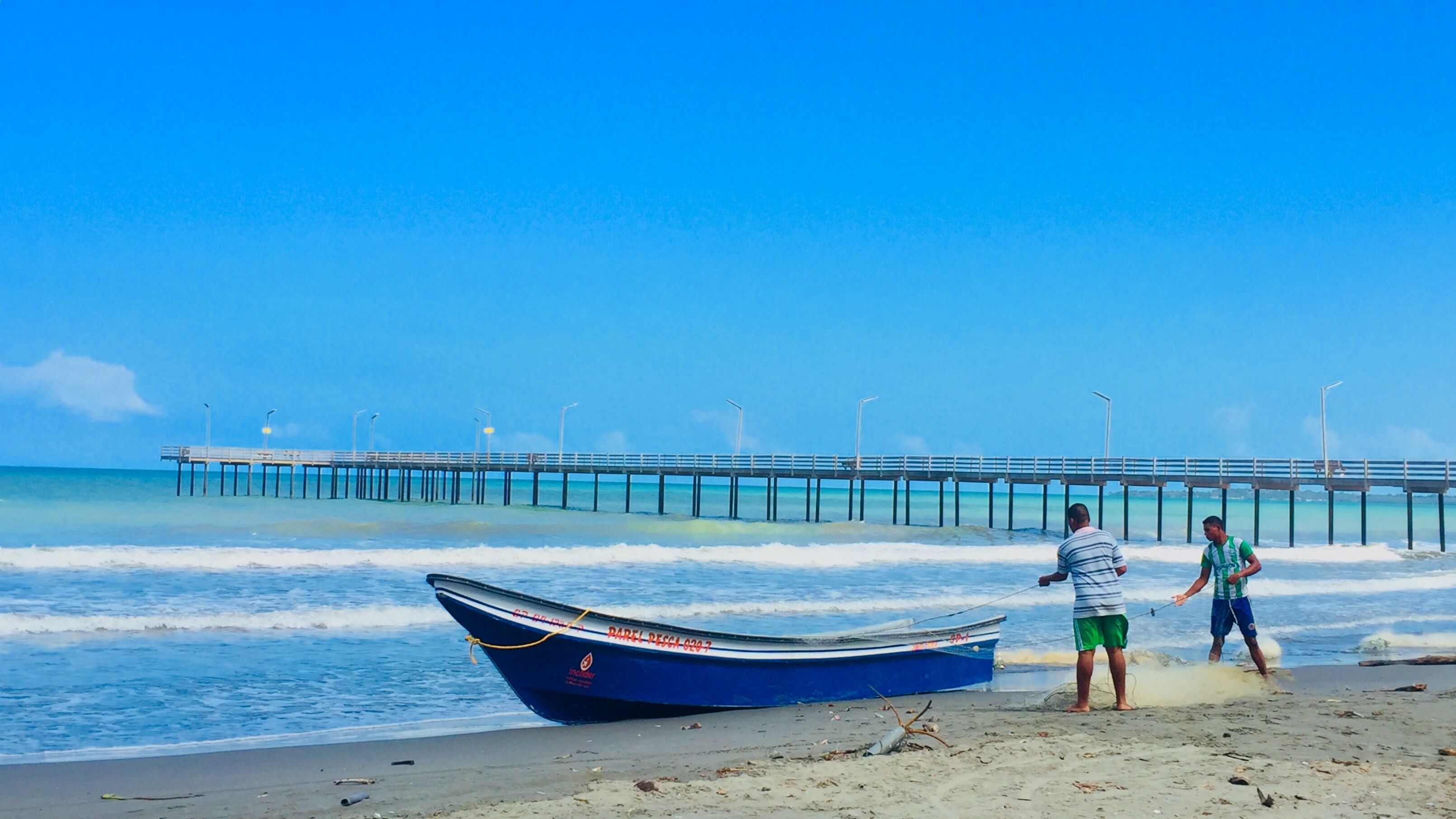 A beach with a pier extending into the ocean, a boat on the sand, and two people near the water.