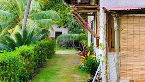 A thatched-roof bungalow with a wooden deck, surrounded by lush greenery and palm trees.