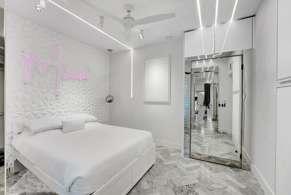 Un dormitorio moderno con una cama, un letrero de neón, un ventilador de techo y un pasillo que conduce a un baño.