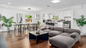 Una sala de estar moderna con un sofá seccional, una mesa de comedor con sillas, una zona de cocina con armarios blancos y plantas en macetas.