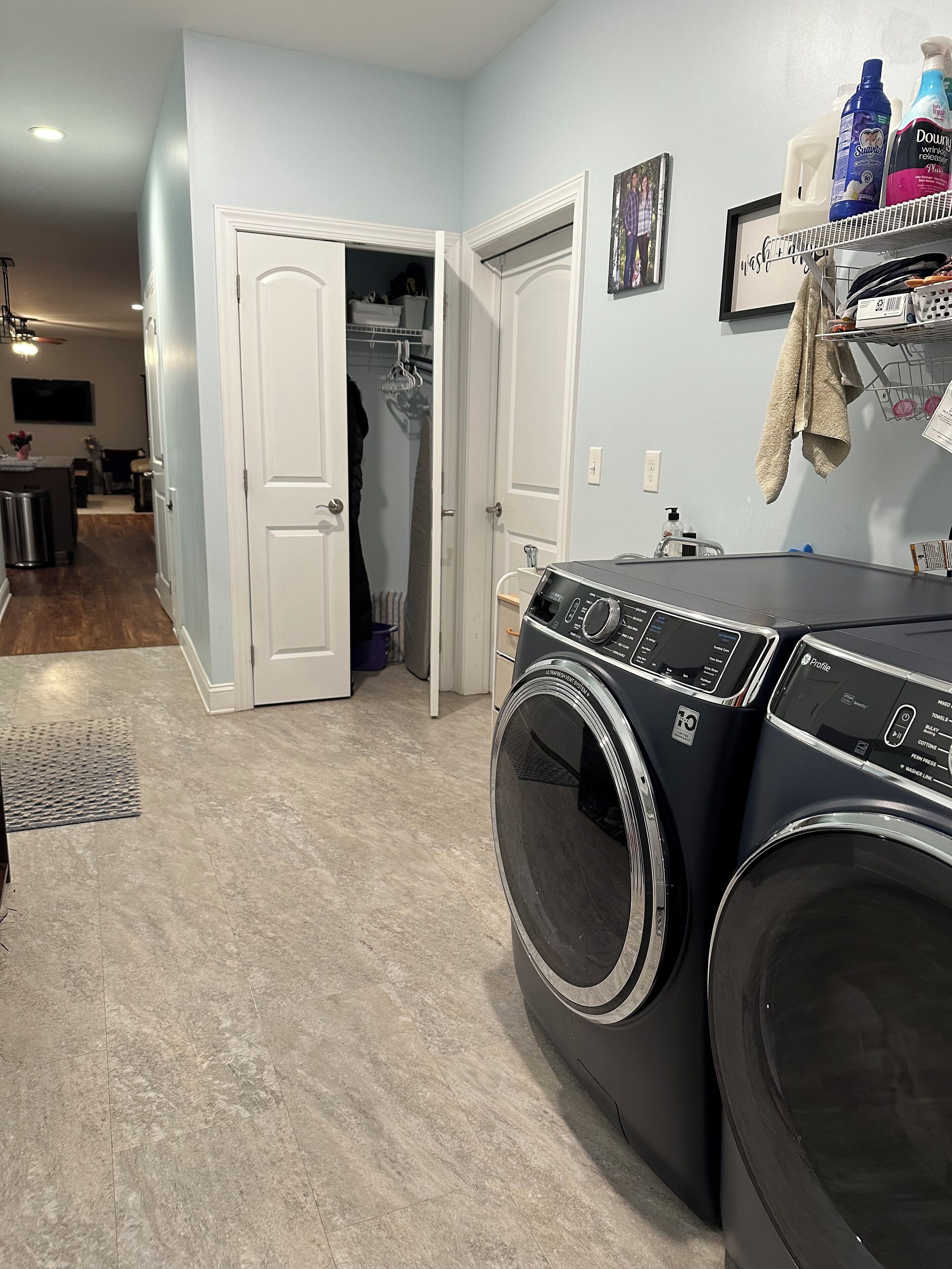 A laundry room with two front-loading washers and dryers, a white door leading to a closet, and a shelf with cleaning supplies.