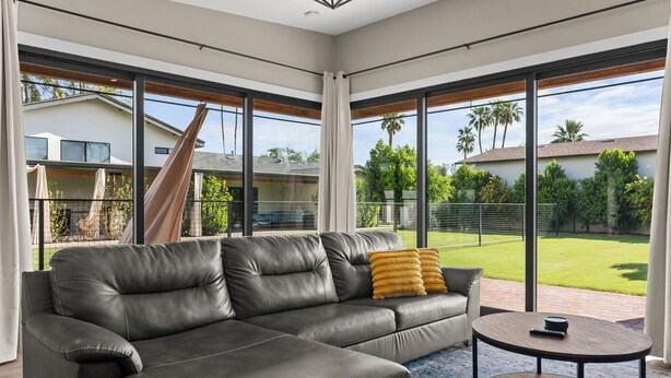 A modern living room with a leather sofa, a round coffee table, and large windows offering a view of a backyard and neighboring houses.