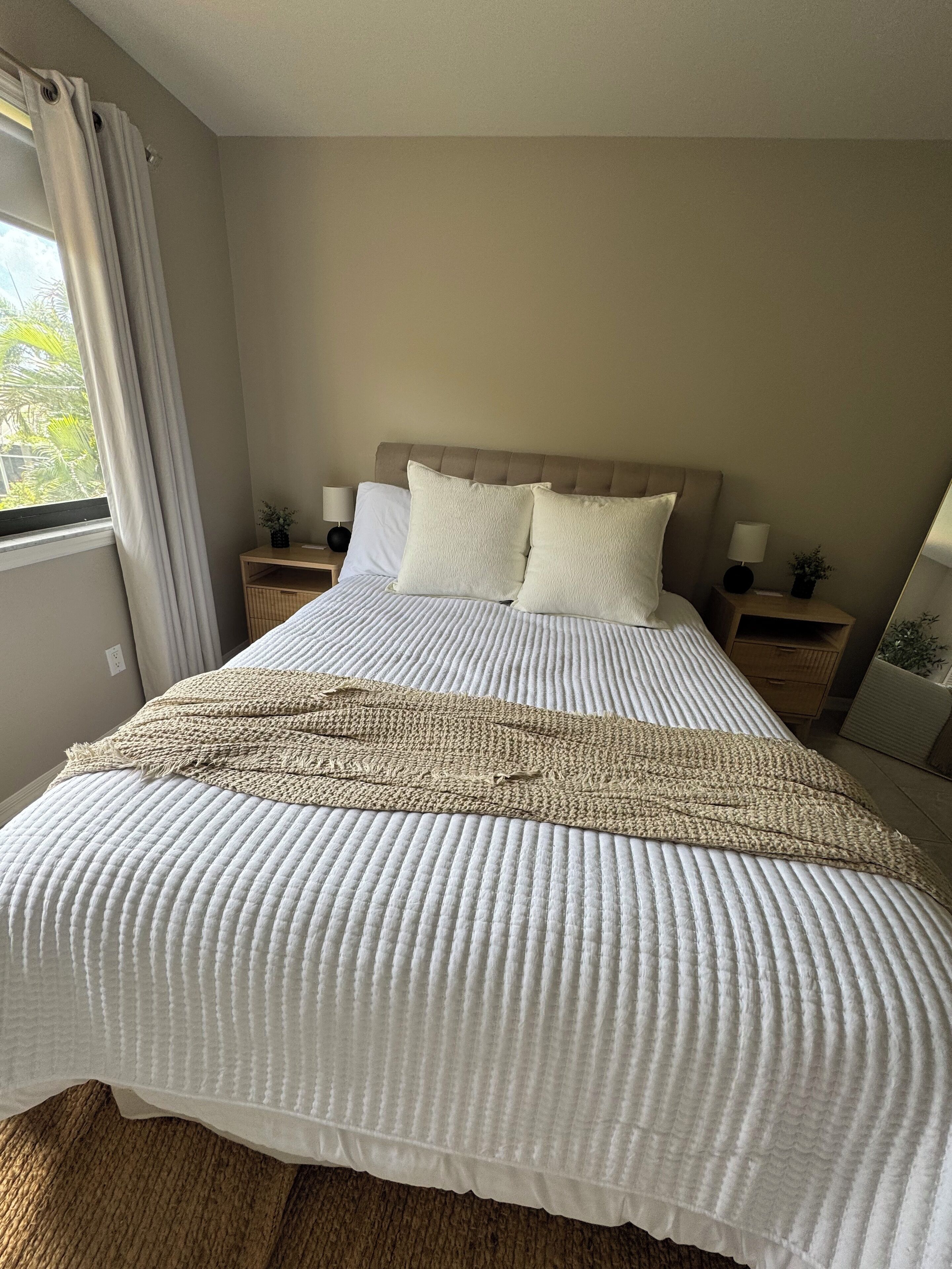 A neatly made bed with a white quilt and a beige throw blanket, two white pillows, and a wooden headboard.