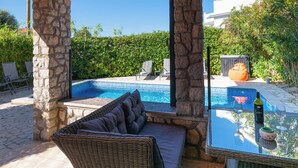 A covered outdoor area with a pool, stone pillars, wicker furniture, and a glass table with wine glasses and a bottle.