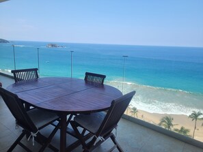 A round wooden table with four chairs, overlooking a beach with clear blue waters and a sandy shore.