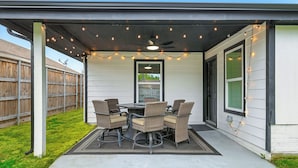 An outdoor patio with a table and chairs, a ceiling fan, string lights, and a view of a wooden fence and house in the background. - Flower Mound Manor - Modern 3BR New Construction (Flower Mound)
