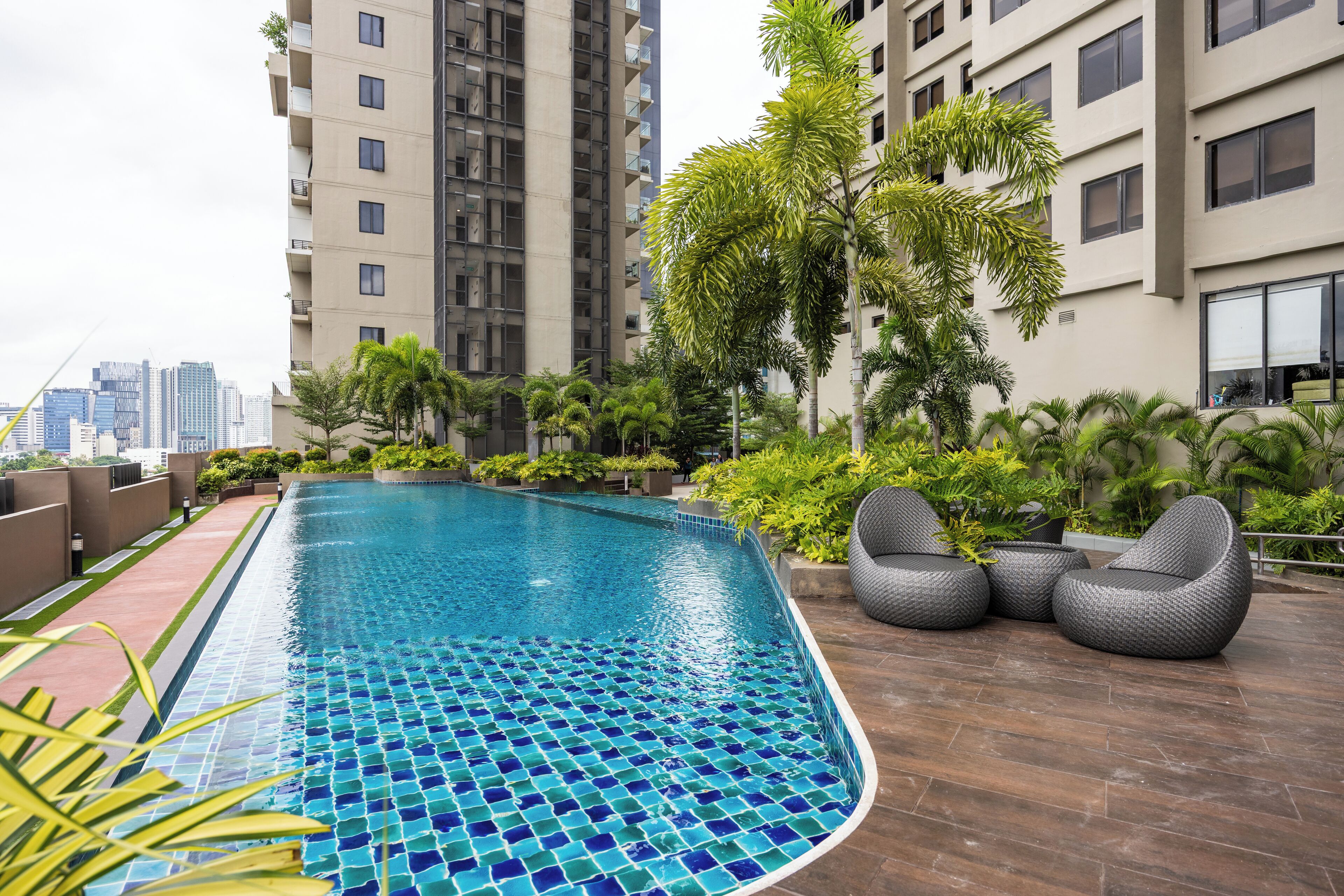 A rooftop swimming pool with tiled blue and green mosaic, surrounded by lush greenery and modern buildings.