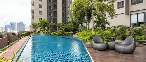 A rooftop swimming pool with tiled blue and green mosaic, surrounded by lush greenery and modern buildings.