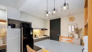 Eine moderne Küche mit weißen Schränken, einem schwarzen Kühlschrank, einer Mikrowelle und einem Spülbecken. Zwei Pendelleuchten, ein Holzstuhl und ein Esstisch sind vorhanden.