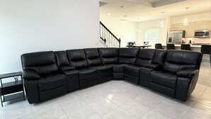 Una sala de estar amplia con un sofá seccional grande de cuero negro, un comedor con mesa y sillas, una cocina con electrodomésticos modernos, y una escalera que conduce a otro nivel.