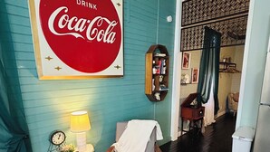 Une pièce aux murs de couleur turquoise, ornée d’une enseigne Coca-Cola, avec une chaise en bois, une petite table sur laquelle se trouve une lampe, et un pouf en osier.