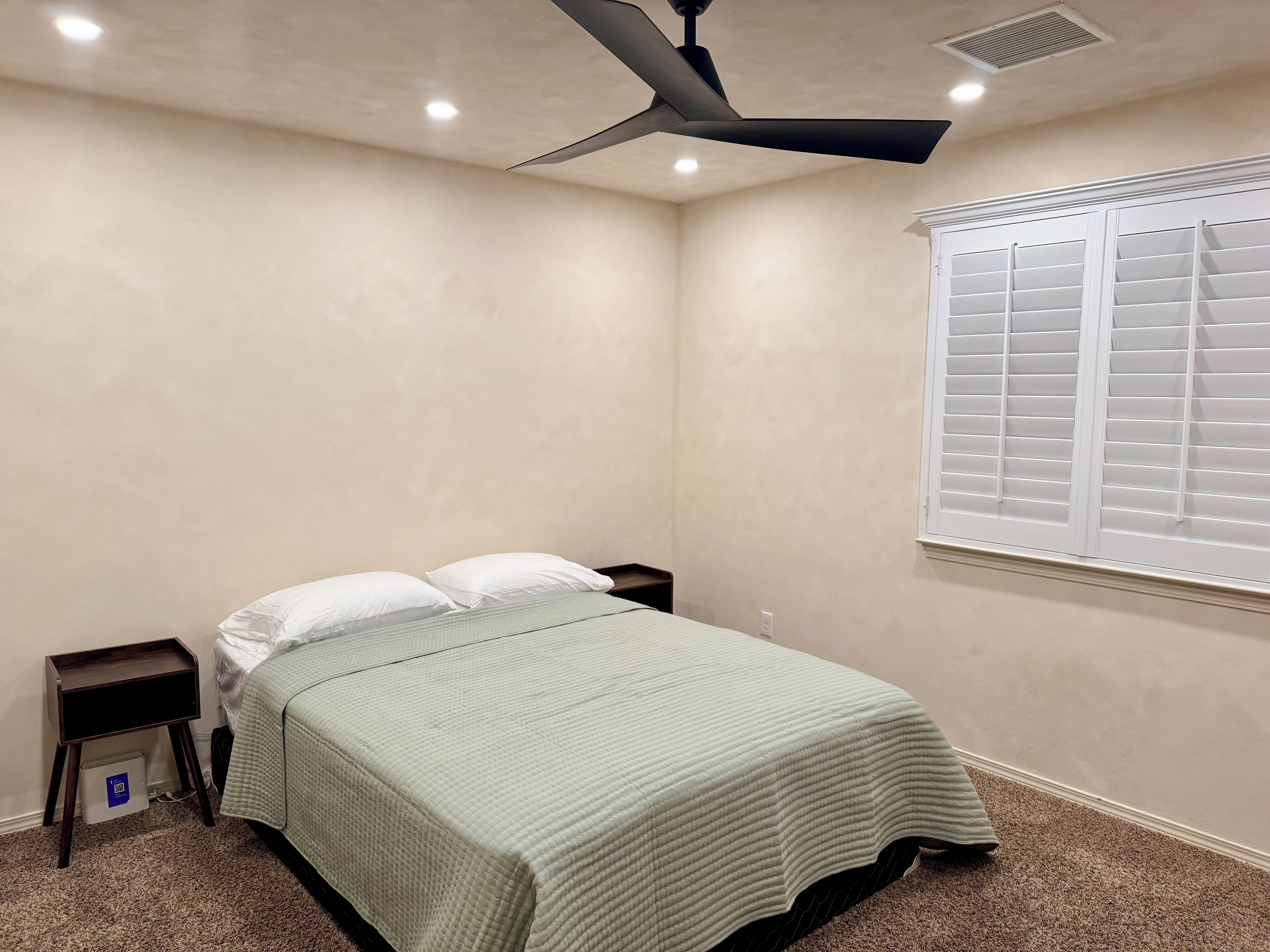 Un dormitorio con cama, mesitas de noche, ventilador de techo, ventana con persianas y piso alfombrado.