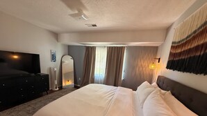 Quarto de hotel com uma cama grande, televisão de tela plana, cômoda, espelho e janela com cortinas.