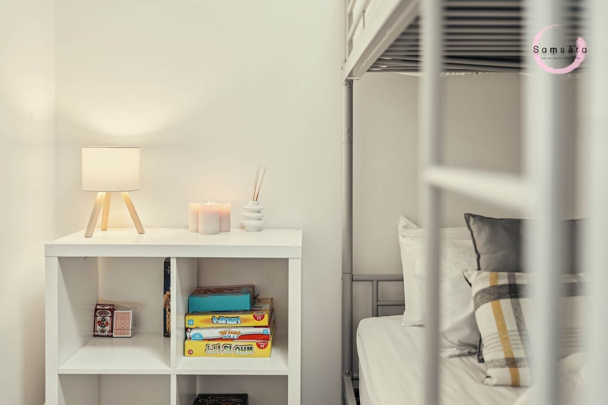 Un dormitorio ordenado con una cama blanca, una mesita de noche con lámpara, velas y una estantería con juegos de mesa.