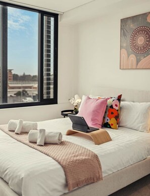 A modern bedroom with a large window, a bed with white linens, a laptop on the bed, and a decorative wall piece. - Saṃsāra - Residence on Flinders (Adelaide)