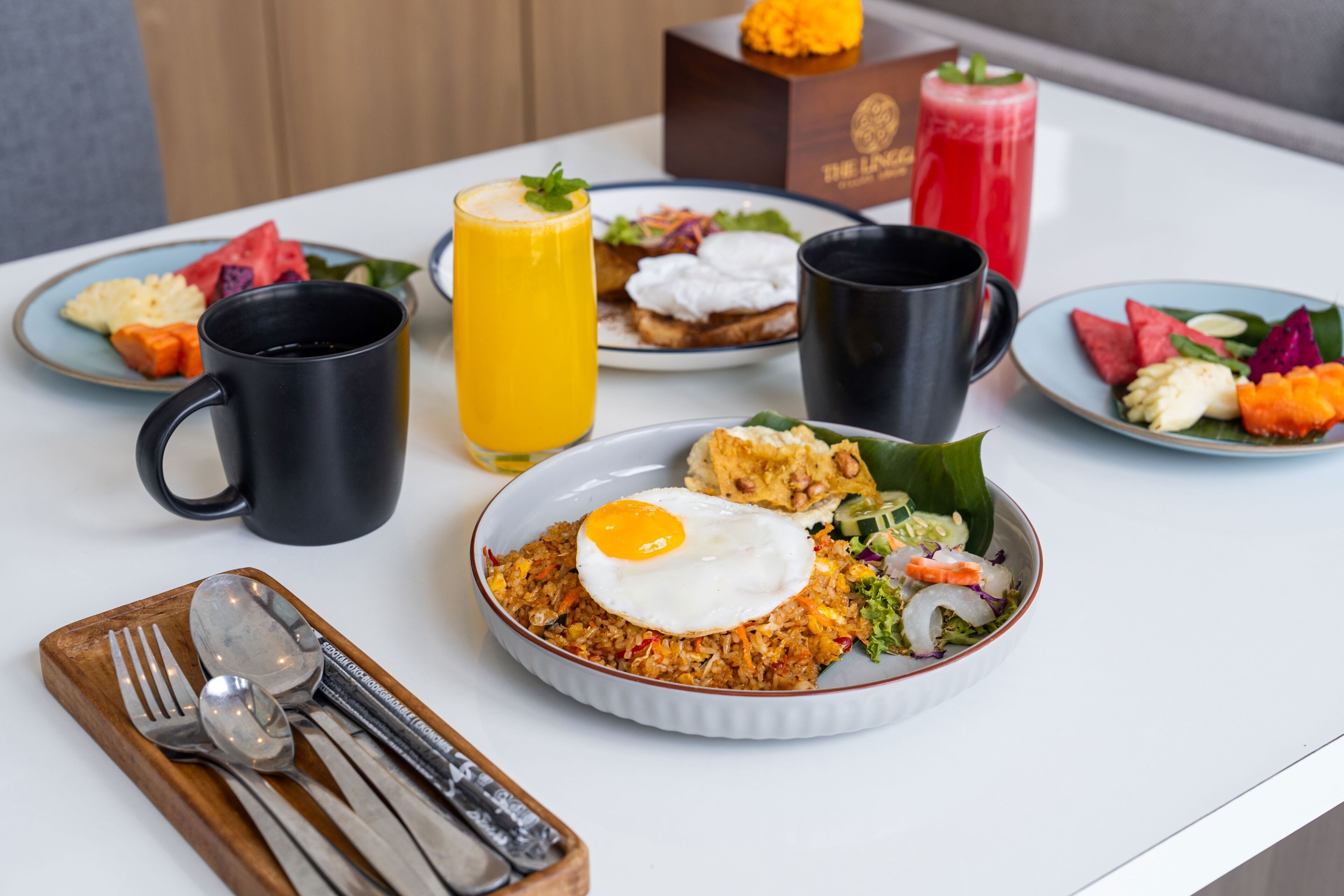 A breakfast setting with a plate of fried rice topped with a fried egg, a bowl of salad, and a plate of fruit. There are also two black mugs and a tall glass of orange juice.
