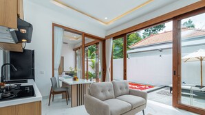 Una cocina moderna con estufa integrada, un comedor con mesa y sillas, un sofá y un ventanal corredizo que da a un patio con un cantero de flores rojas.