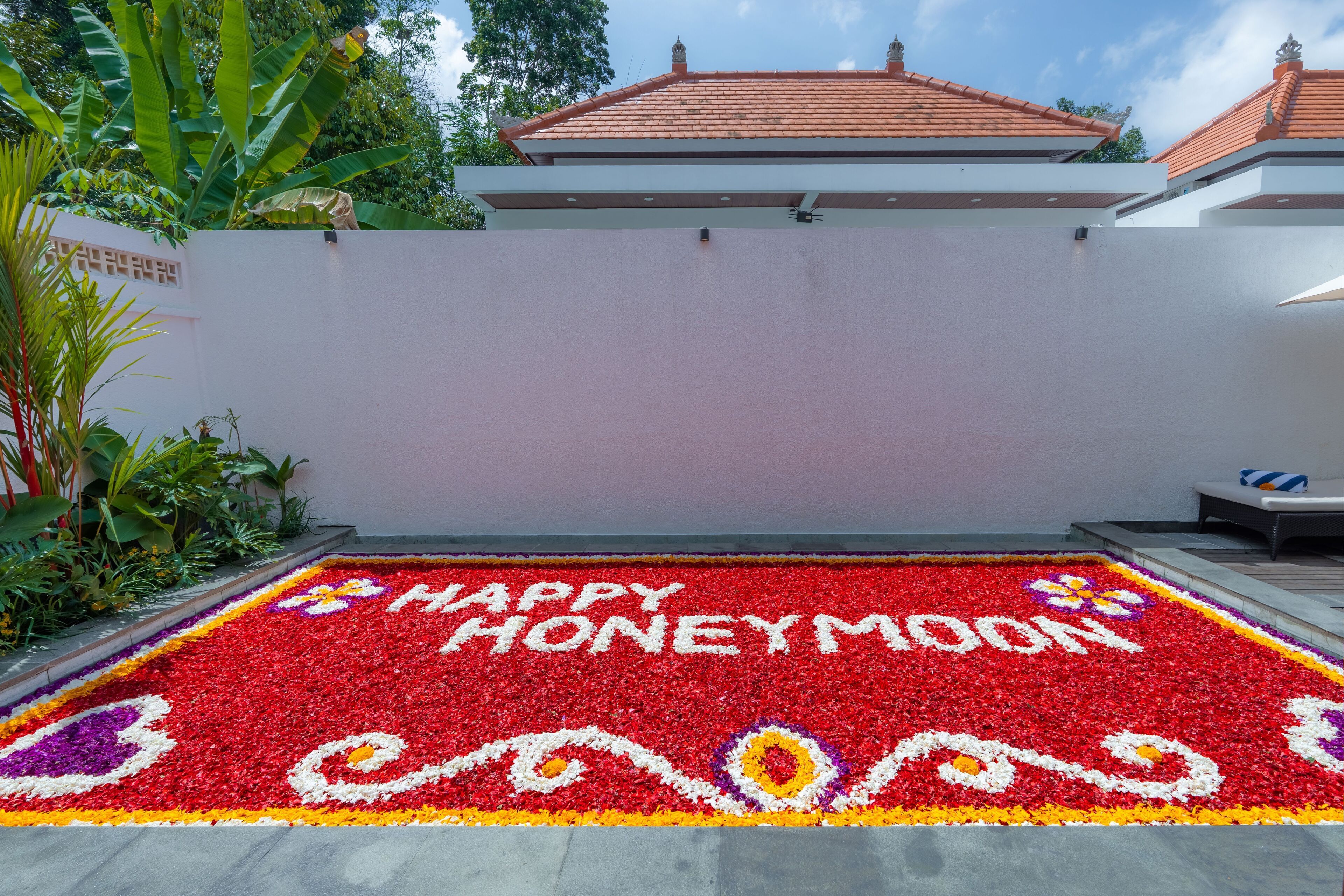 Un jardí amb un conjunt de flors vermelles formant la frase “HAPPY HONEYMOON” i una paret blanca amb un dibuix decoratiu.