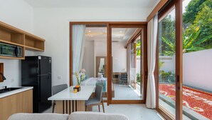 Una cocina moderna con un refrigerador negro, armarios de madera y un microondas. Hay un comedor con una mesa y sillas blancas, y una vista de un jardín a través de una puerta de vidrio.