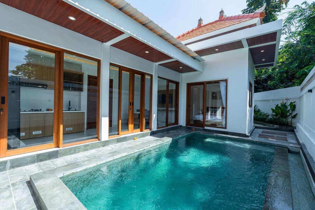 A modern house with a swimming pool, large sliding glass doors, and a kitchen visible through the glass.
