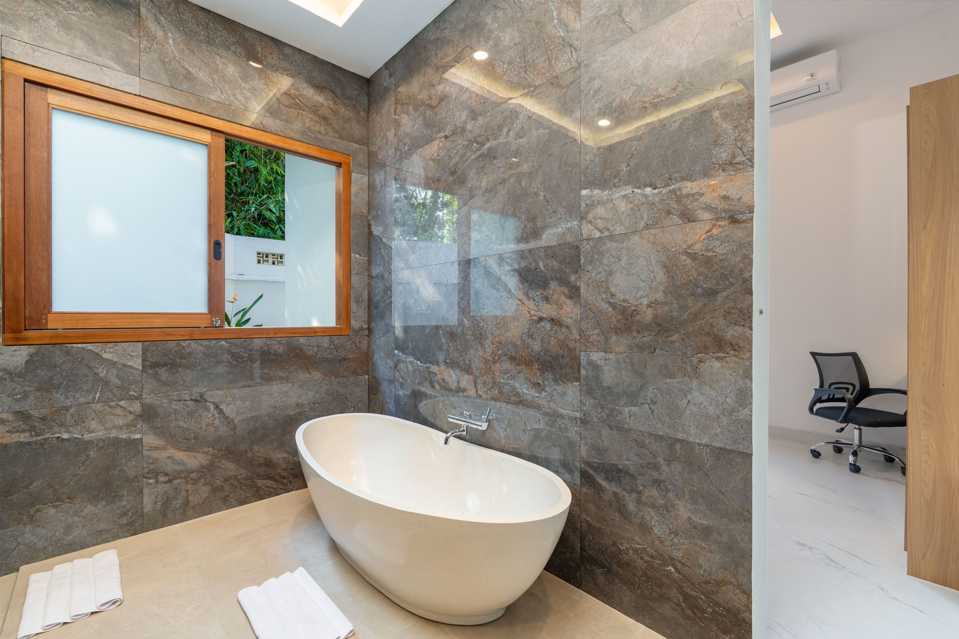 Une salle de bain moderne avec une baignoire sur pieds, des murs en marbre, un miroir encadré en bois et un petit fauteuil de bureau placé dans un coin.