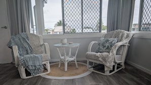 A room with two white wicker chairs, a small round table, and a view through a window with a metal screen.