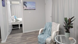 A room with a white chair, a small table with a plant, a painting on the wall, and a bed with a blue and white patterned blanket.