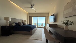 Ett modernt hotellrum med en stor säng, ett skrivbord, en stol, en takfläkt och utsikt över havet genom balkongdörrarna.