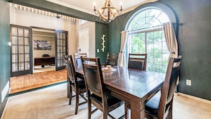A dining area with a wooden table, chairs, and a chandelier. There is a window with curtains and a view of greenery outside. - Spacious Centennial 4BR with Pool Table and Patio (Centennial)
