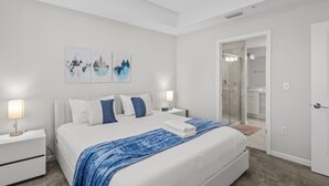 A modern bedroom with a large bed, white walls, and artwork. There is a bathroom visible through a glass door. - Condo 2bd/2bh 15 min from parks Storey Lake (Kissimmee)
