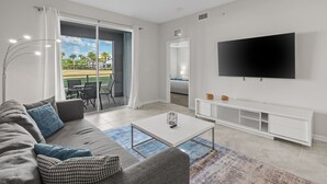 A modern living room with a grey sofa, a flat-screen TV, and a balcony with a view of palm trees. - Condo 2bd/2bh 15 min from parks Storey Lake (Kissimmee)