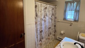 Un baño con puerta de madera, cortina de ducha con diseños de París, lavamanos blanco con grifo negro y una ventana con cortinas traslúcidas.