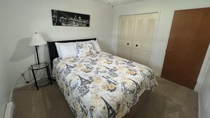 Un dormitorio con cama, ventilador de techo, mesita de noche, lámpara, armario y un cuadro colgado en la pared.