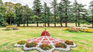 A park with a central flower arrangement, benches, and tall trees. - Varis Haven by Interhome (Forres)