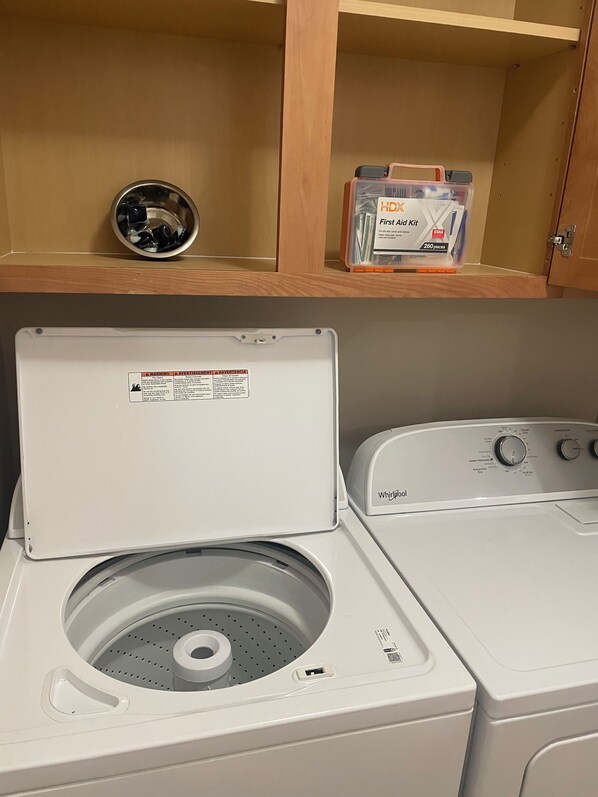 A front-loading washer and dryer in a laundry room with a shelf above containing a first aid kit and a metal bowl.