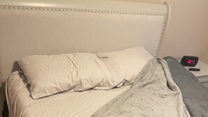Un lit avec une tête de lit blanche, une couverture grise et une horloge numérique affichant 02h27.