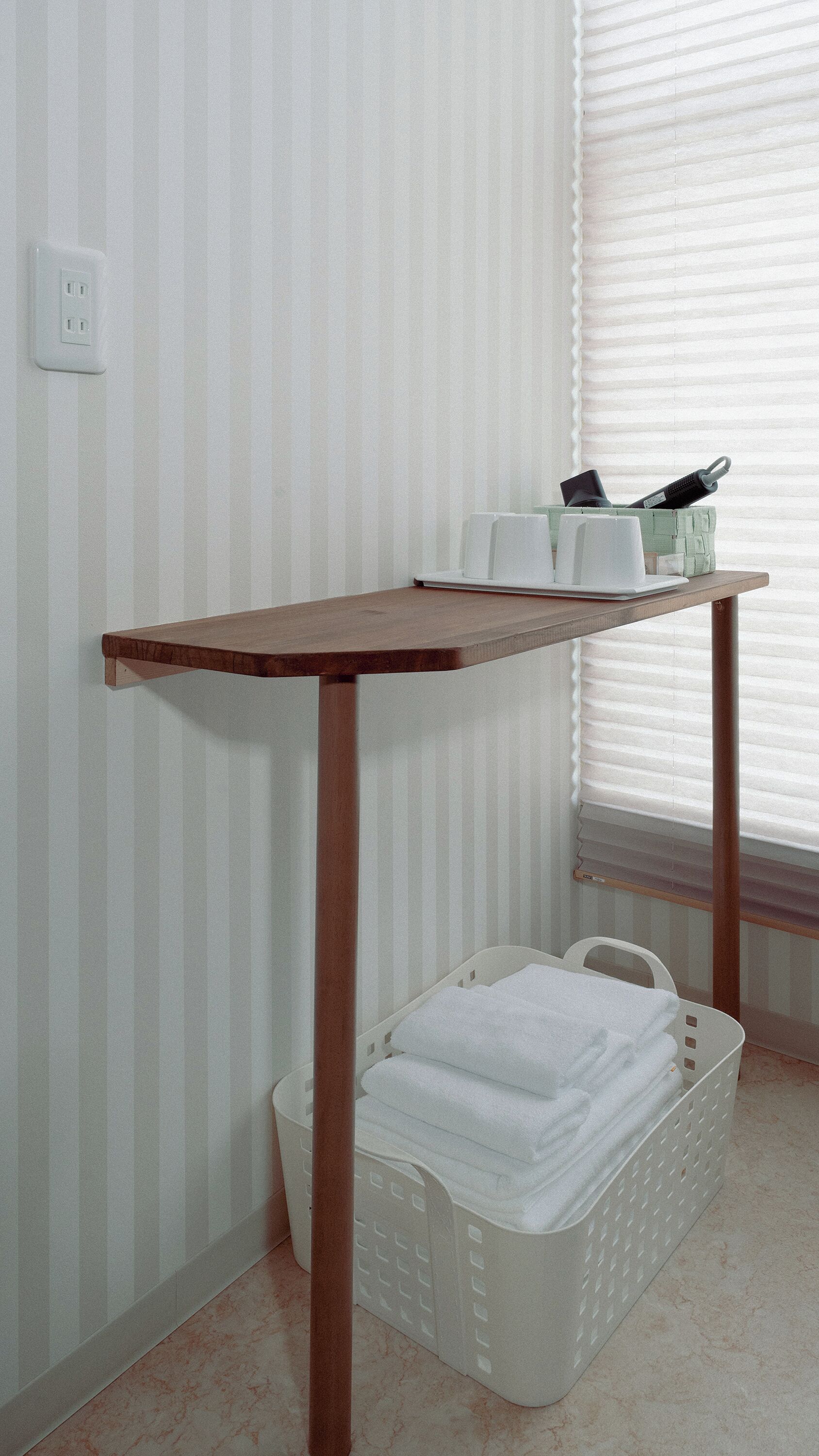A corner with a wooden table, a white basket with towels, and a window with blinds.