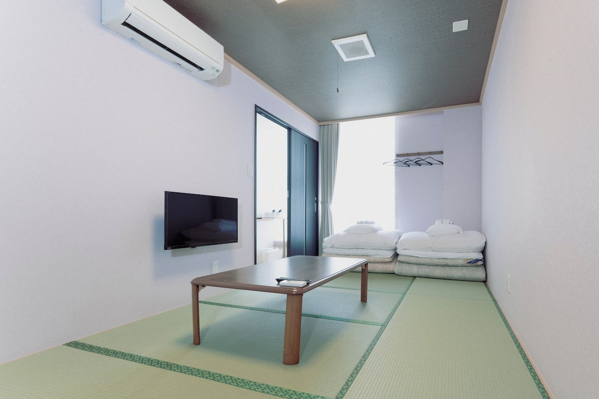 A room with a flat-screen TV, a small table, tatami mats, a sliding door, and a stack of towels.