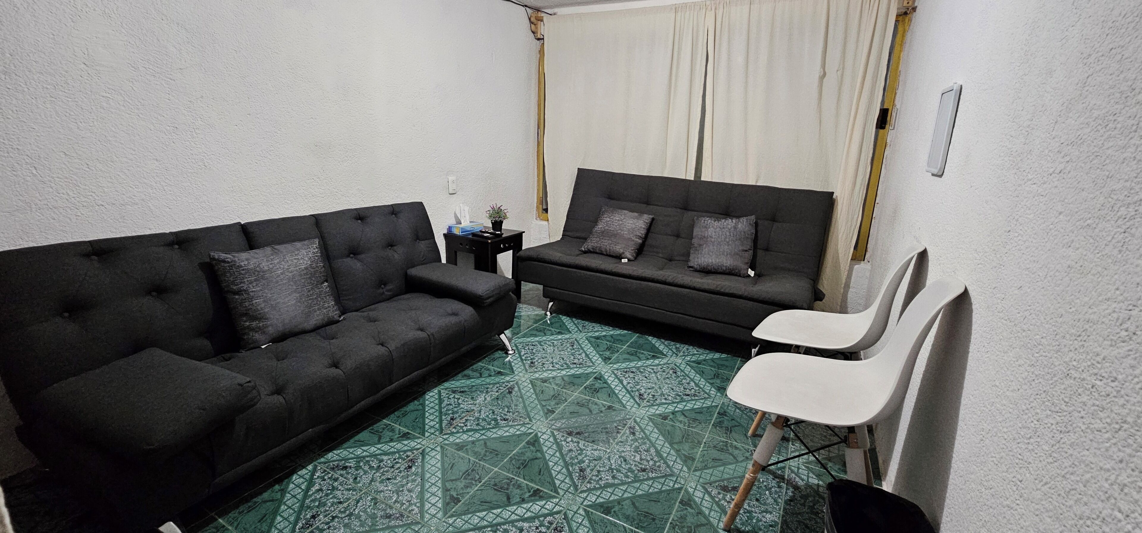 Uma sala de estar pequena com dois sofás pretos, uma poltrona branca e um chão com padrão verde.
