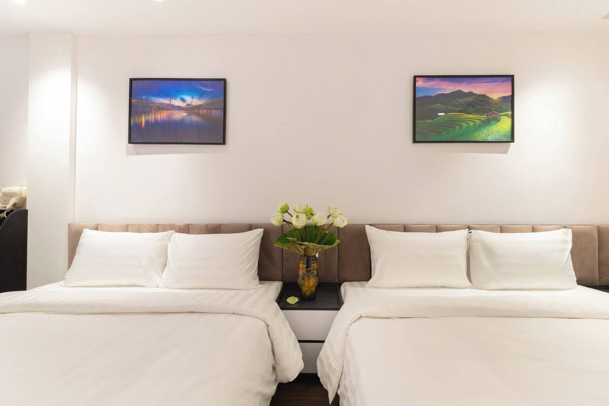 A hotel room with two beds, a nightstand with a vase of flowers, and two framed pictures on the wall.