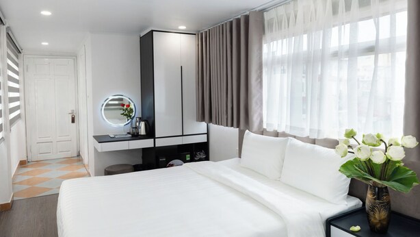 A modern hotel room with a large bed, a vanity with a mirror, a small table with a vase of flowers, and a window with sheer curtains. - Better Home Hanoi (Hanoi)