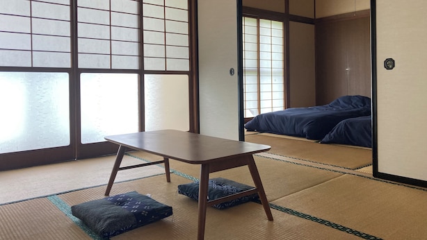 Ein traditioneller japanischer Raum mit Tatami-Bodenbelag, einem niedrigen Holztisch und einem Bett mit dunkelblauer Bettdecke.