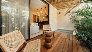Eine moderne Außenpatio mit Holzboden, einer Sitzecke mit gewebten Stühlen, einem kleinen Holztisch und einer Glastür, die zu einem Innenbereich mit Küche und Essbereich führt.