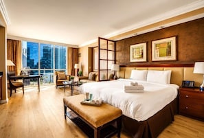 A hotel room with a large bed, a desk, a chair, a bench, and a view of the city.