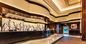 A modern hotel lobby with a curved reception desk, a large wall with a red floral pattern, and a framed artwork on the wall.