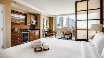 A hotel room with a large bed, a kitchenette, a dining area, and a view of the city.