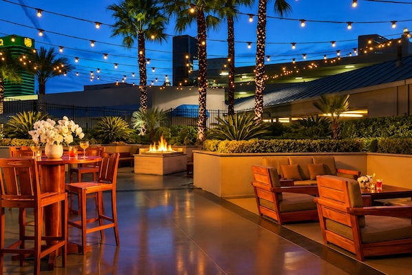An outdoor patio with wooden furniture, a fire feature, and string lights against a night sky.