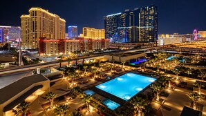 A hotel with multiple swimming pools, a large urban skyline in the background, and a well-lit outdoor area with palm trees.
