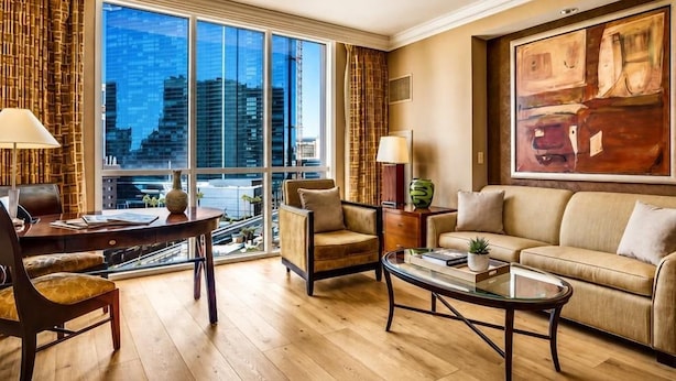 A modern hotel room with a large window, a sofa, a coffee table, and a desk with a lamp.
