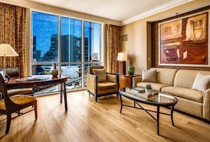 A modern hotel room with a large window, a sofa, a coffee table, and a desk with a lamp.